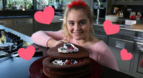 A girl wearing a red love-heart headband smiles next to a chocolate biscuit jewel box covered in fruit and sweets.