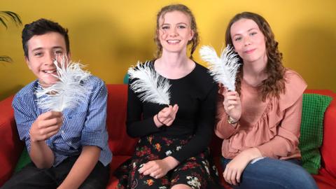 Three members of hetty feather cast sitting on a sofa holding feathers.