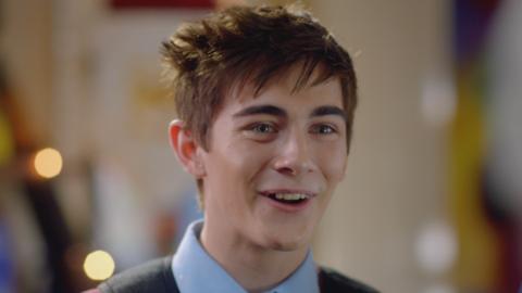 A boy with short brown hair smiles with lights in the background.