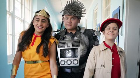 Three teenagers dressed up in fancy dress walking down a school hallway.