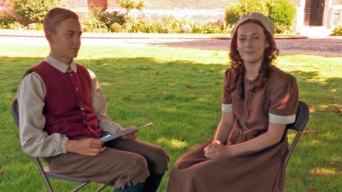 A boy and girl in Victorian outfits sat in a courtyard. (Cast of Hetty Feather on set).