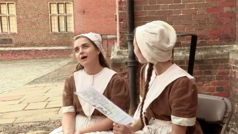 Two girls in Victorian outfits sat in a courtyard. (Cast of Hetty Feather on set).