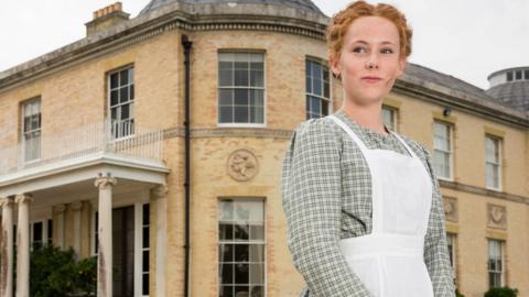 A girl in victorian servant dress standing in front of a grand house.