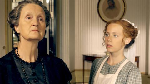 A girl in a victorian servant uniform looking up at an elderly lady who looks cross.