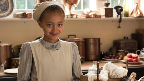 A young girl wearing victorian servant clothes in a kitchen.