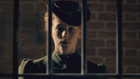 A girl in Victorian dress standing behind jail bars.