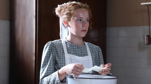 A girl reading a piece of paper in a kitchen looking shocked.