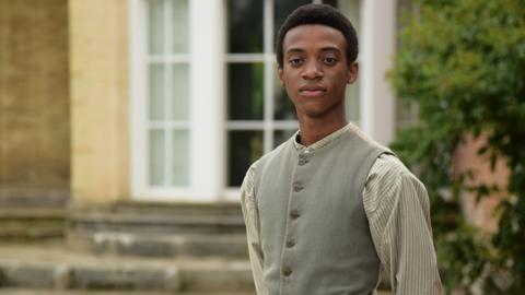 A boy wearing Victorian clothing stands in a garden.