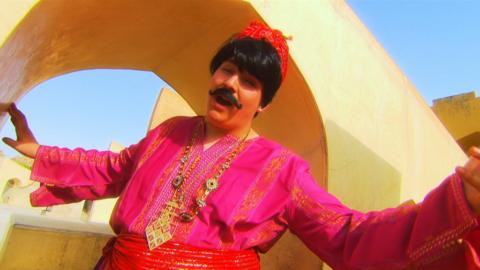 A man wearing a fake moustache, wig, fez and red robe gestures broadly while standing in a doorway.