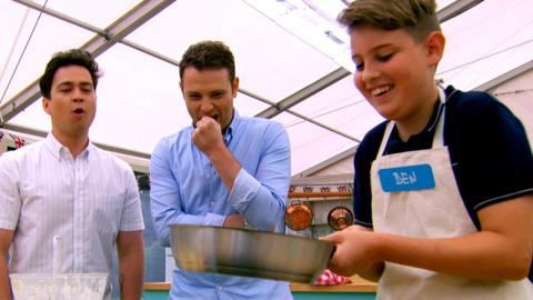 A boy gets ready to flip a pancake with a frying pan while two presenters looking on (Sam and Mark).