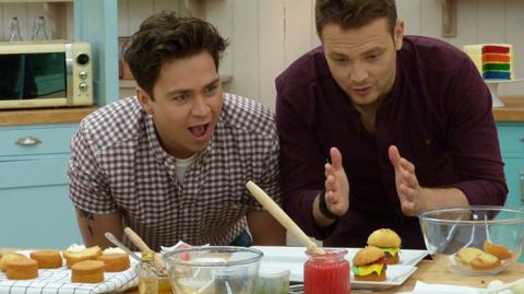 Sam and Mark lean over a counter in the Bake Off tent admiring some cakes.