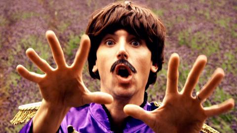 Ed, dressed as a Beatle, holds up his hands in shock, dressed in purple.