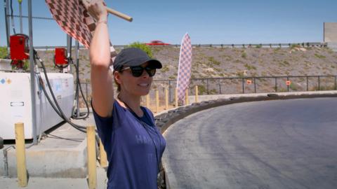 A woman wearing a cap and sunglasses holds up a checkered race flag to signal the start of a go kart race.
