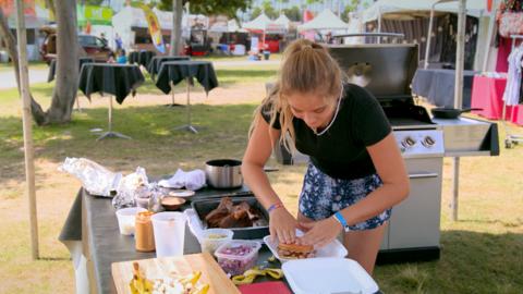 A girl prepares a burger surrounded by barbecue equipment.