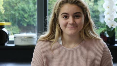 Tilly, in a pink jumper, sits in her kitchen smiling.