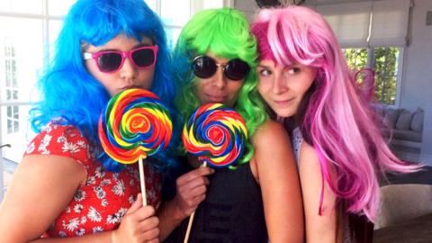 Three women pose with brightly coloured wigs and lollipops.