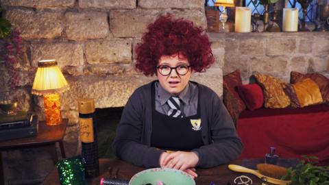 A girl in school uniform at her desk wearing a curly red wig. Maud from The Worst Witch.