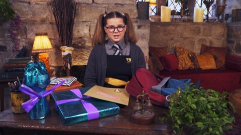 A girl sat at her desk with lots of items ready to pack into a bag. Maud from The Worst Witch.