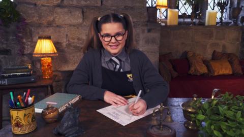 A girl in school uniform sat at her desk writing. Maud from The Worst Witch.