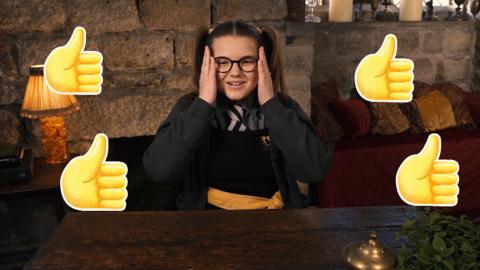 A girl in school uniform at her desk with her hands to her face. Thumbs up emojis surround her.