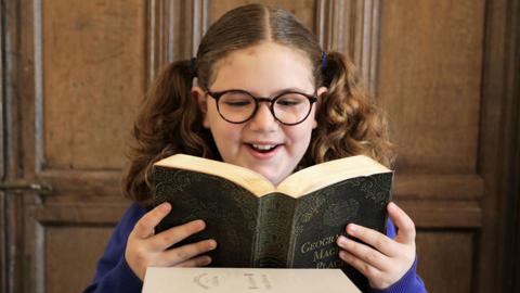 A girl with curly hear in bunches, wearing glasses and looking excited reading an old looking book (Maude)