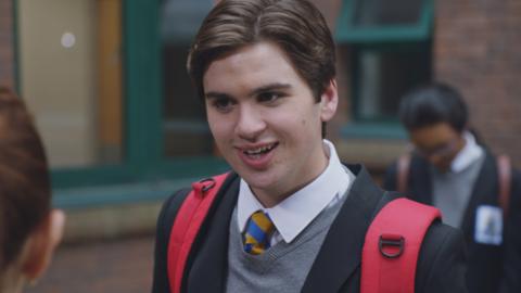 A teen boy wearing a school uniform and a red backpack smiles at a girl in the school playground. (Craig).