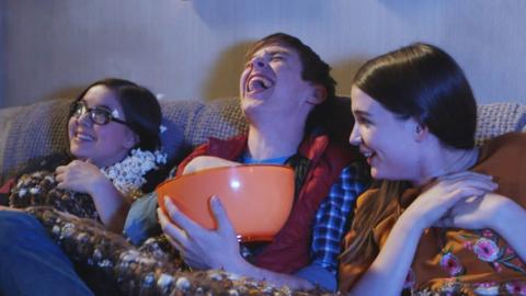 Millie, Leo and Lauren sat on the sofa laughing with Miliie covered in popcorn and Leo holding a bowl.