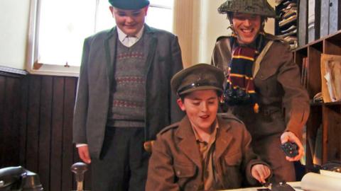 Ed stands with two boys, all are dressed in 1940s style WW2 uniform. They are looking at a morse code 'straight key'.