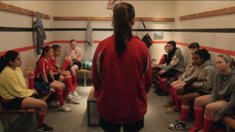 Marnie is standing in the football changing rooms. The other players are sitting on benches around her.