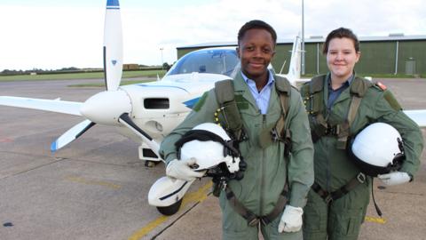 A boy and a girl holding helmets stood in front of a small plane.