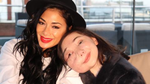 A girl sat on a sofa cuddling a woman, both smiling. Nikki Lilly and Nicole Scherzinger.