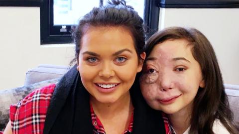 Two girls sat on a sofa smiling, Nikki Lilly and Scarlett Moffatt.