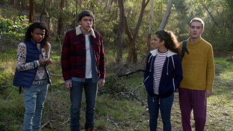 Three boys and a girl looking surprised, standing in a wooded clearing.