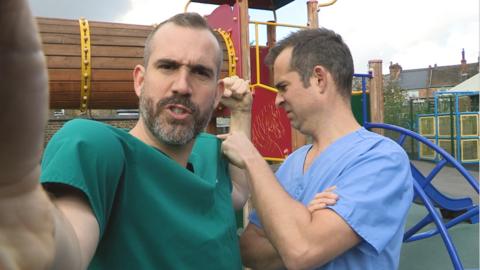 Dr Xand flexing his muscles while Dr Chris pokes at them. They are both standing in a playground.