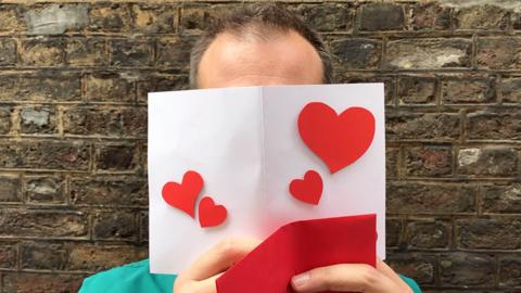 Dr Xand holding a white card with red love hearts on it. He is standing in front of a brick wall.