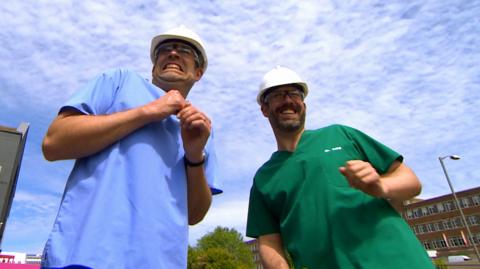 Two men in doctor's scrubs also wearing hard hards looking surprised.