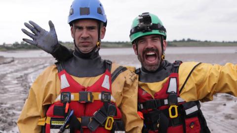 Two men wearing yellow wader suits and blue helmets on a very muddy beach looking happy. Dr. Chris and Dr. Xand from Operation Ouch.