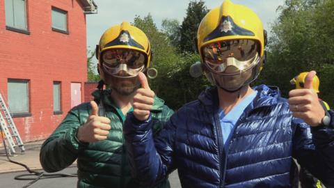 Two men wearing firemans helmets covering their face while making a thumbs up gesture.