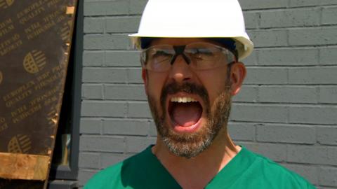 A man wearing a hard hat and green scrubs is shouting.