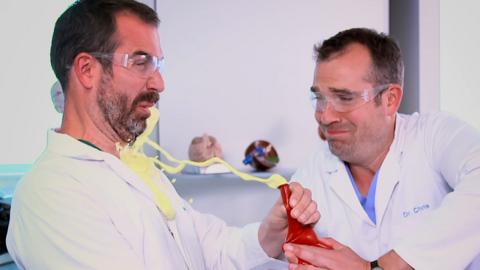 Two doctors wearing lab coats and one is squeezing out yellow foam from a balloon into the others face.