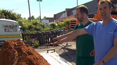 Two men wearing scrubs are pointing at a pile of sand being used as a model of the brain.