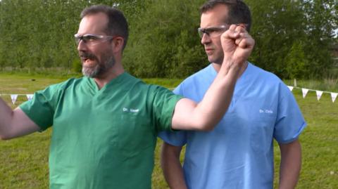 Two doctors in scrubs and goggles, stand in a field cheering.