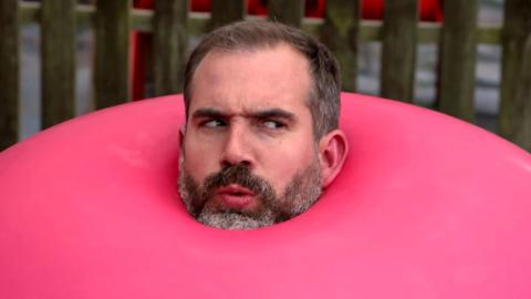 A man with a beard looks confused as his head appears to be sticking out of a giant inflatable red ball, Dr Xand from Opertion Ouch.