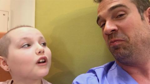 A man in blue scrubs sat next to a young girl with short hair who is smiling, Dr Chris and Ruby from Operation Ouch.