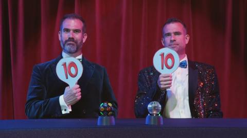 Two men sat behind a desk in sparkly suits holding up paddles with 10 on them.