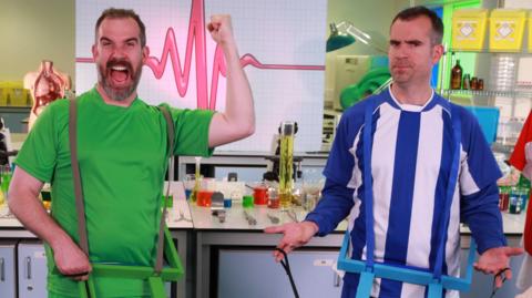 Two doctors in the lab wearing football kits while one of them cheers, Dr Xand and Dr Chris from Operation Ouch.