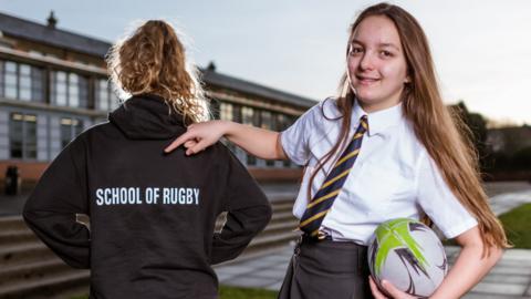 A girl with a rugby ball pointing at another girl's back that says 'Rugby Team'