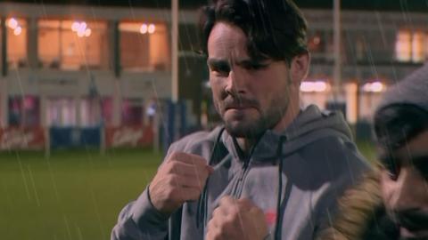 Man on a rugby pitch in a grey hoodie with two fists up like he's in a fight.