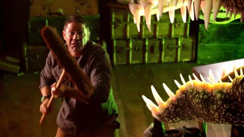 Steve Backshall using a broom to scare away a dinosaur.