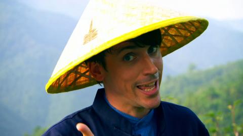 A man wearing traditional Chinese working clothes and a woven hat points to the camera on a hilly tea plantation.